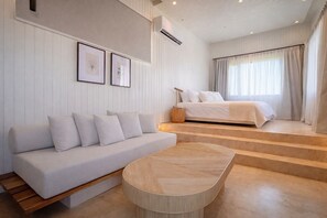 A modern bedroom with a bed, a sofa, and a small table. There are stairs leading to the bed, and the room has a minimalist design with white walls and a wooden floor.