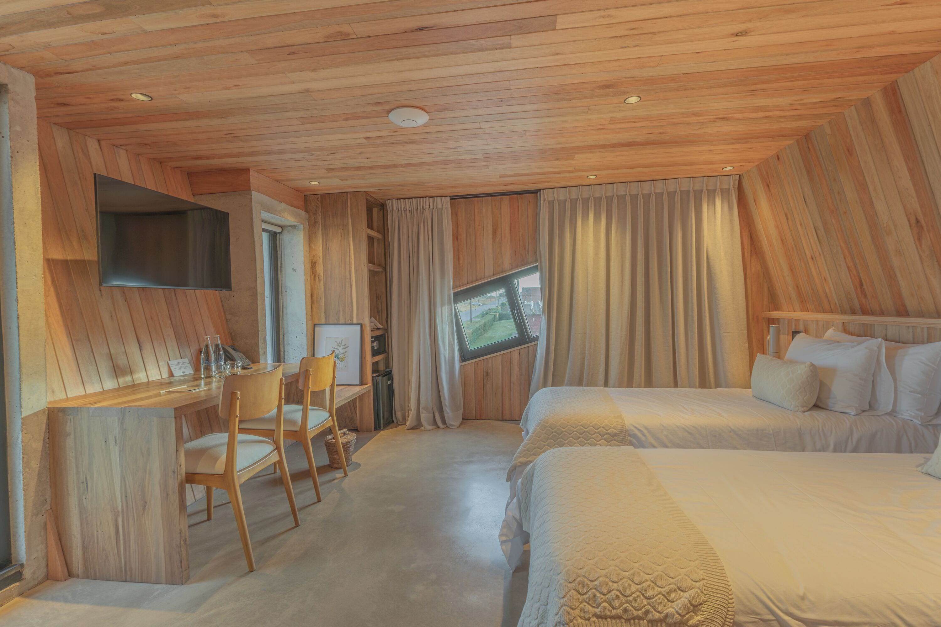 A hotel room with a wooden ceiling, a desk with chairs, a bed, and a television mounted on the wall.
