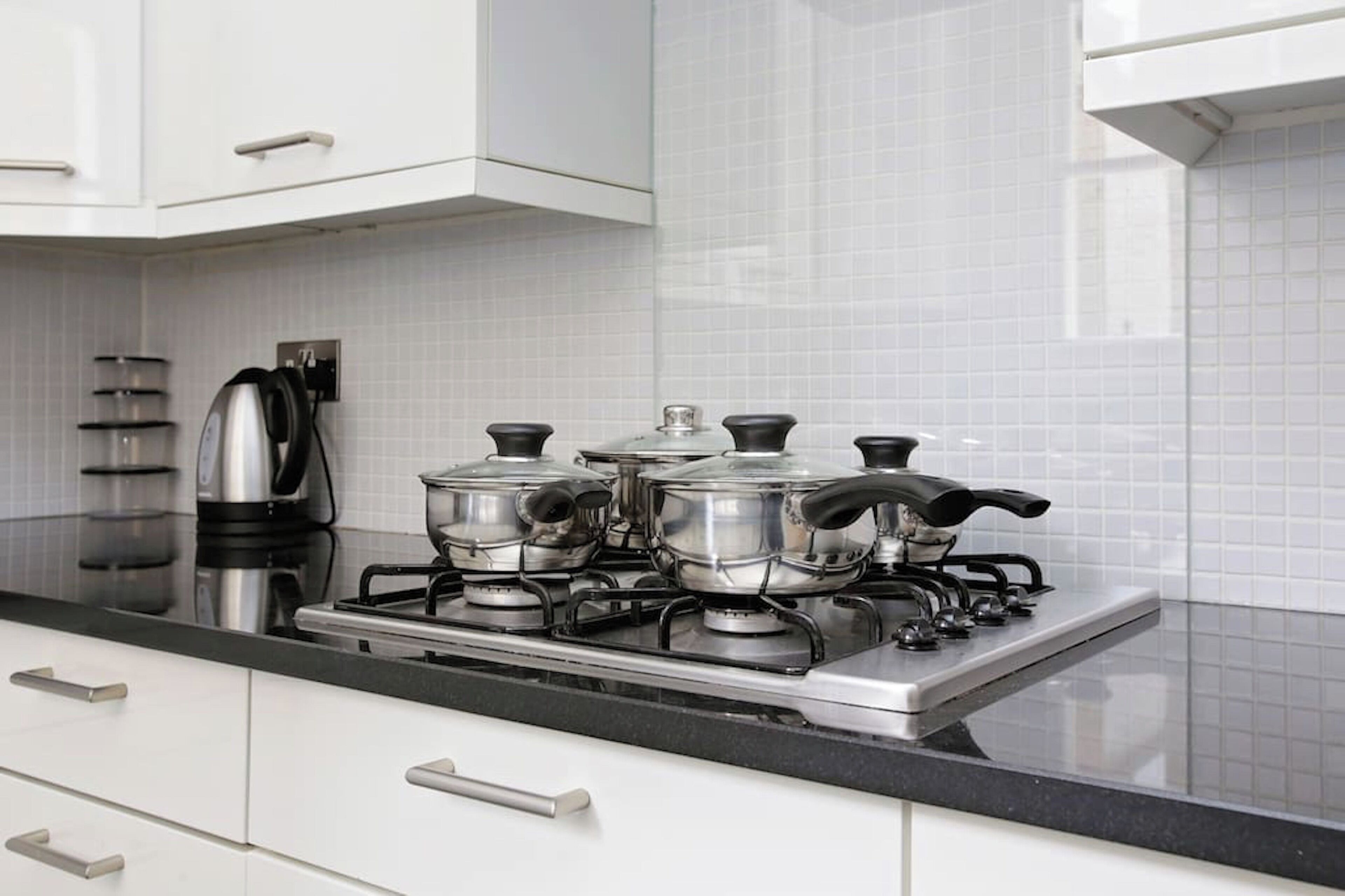 Cucina con mobili bianchi, piano di lavoro nero e fornello a gas con pentole e padelle.