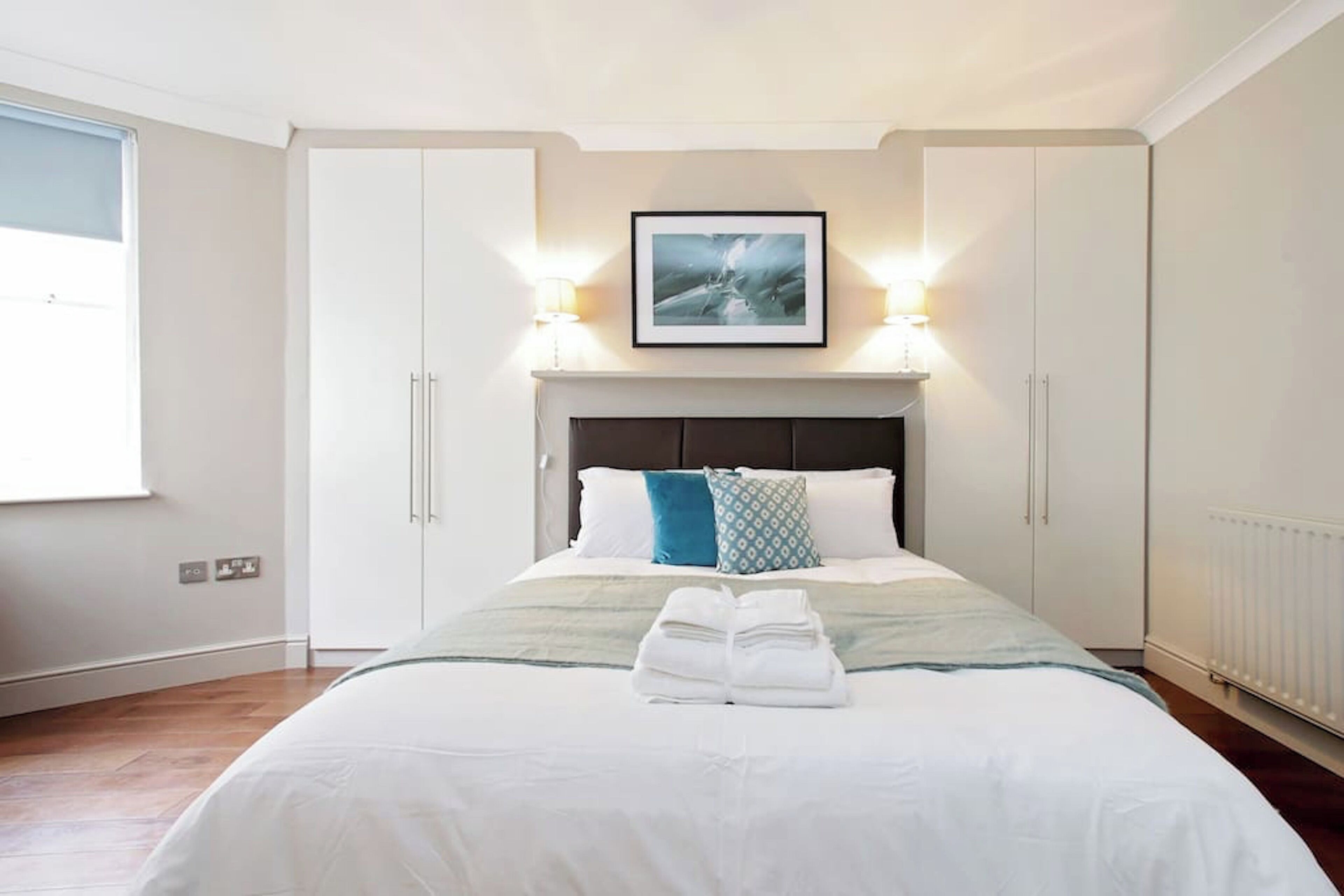 A neatly made bed with a folded towel, a framed picture above the headboard, and two wall-mounted lamps.