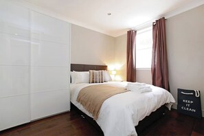 Ein ordentlich bezogenes Bett mit weißen Laken und einer beigen Tagesdecke, eine Nachttischlampe und ein Schild mit der Aufschrift 'KEEP IT CLEAN'.