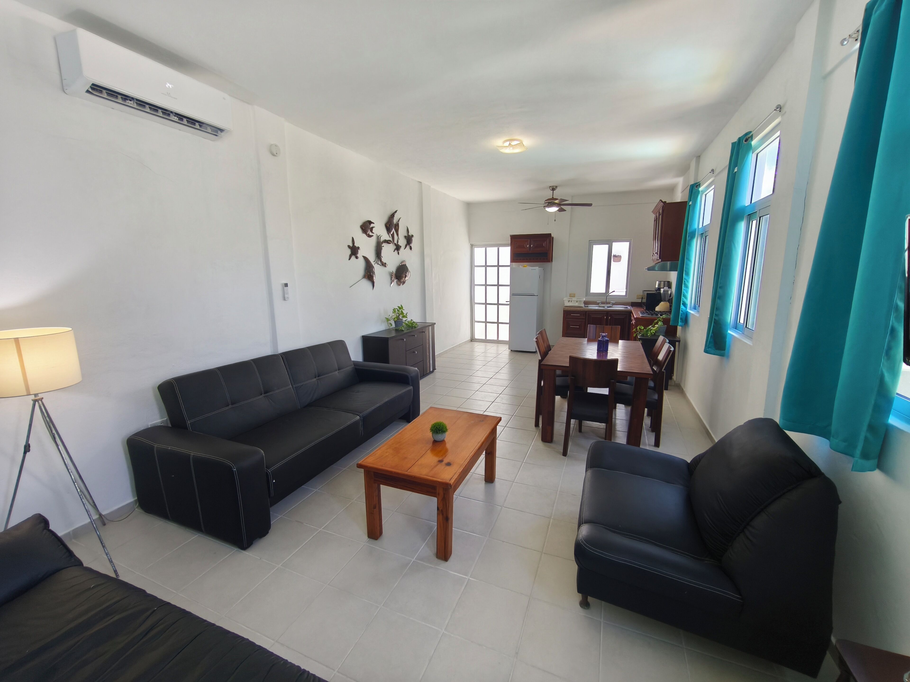 A living room with a sofa, coffee table, and dining table. There is a kitchen area in the background with cabinets and a refrigerator.