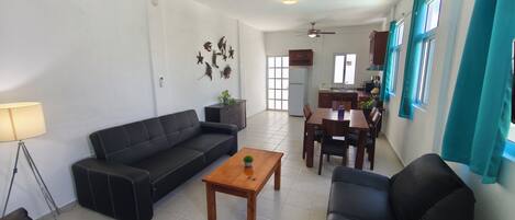 A living room with a sofa, coffee table, and dining table. There is a kitchen area in the background with cabinets and a refrigerator.