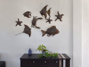 A wall with a collection of metal fish and sea turtle decorations. Below, a black cabinet holds a blue bottle and a potted plant. - Azul Mar Apartment 2 (Puerto Morelos)