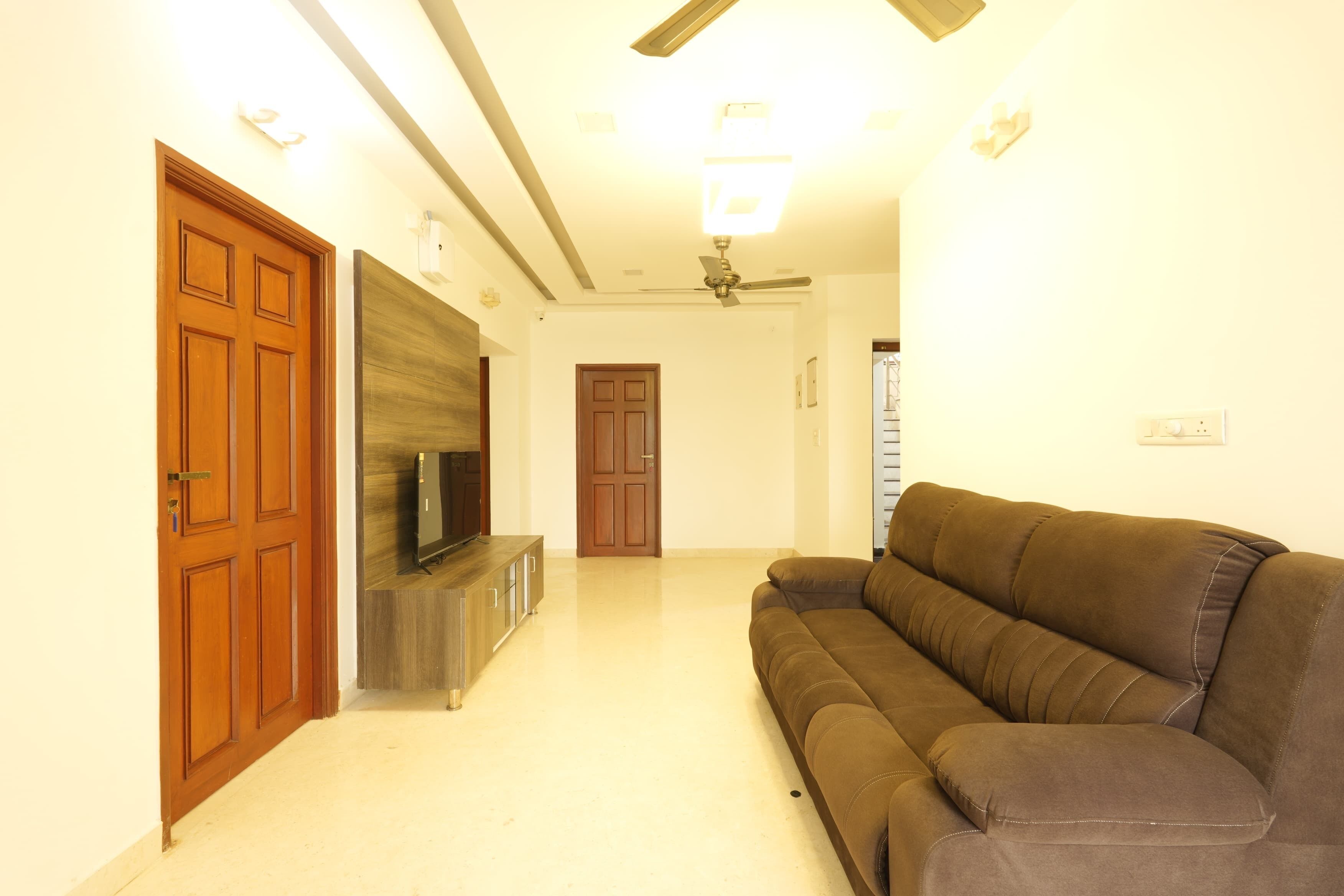 A living room with a brown sofa, wooden doors, a TV on a wooden cabinet, and a ceiling fan.