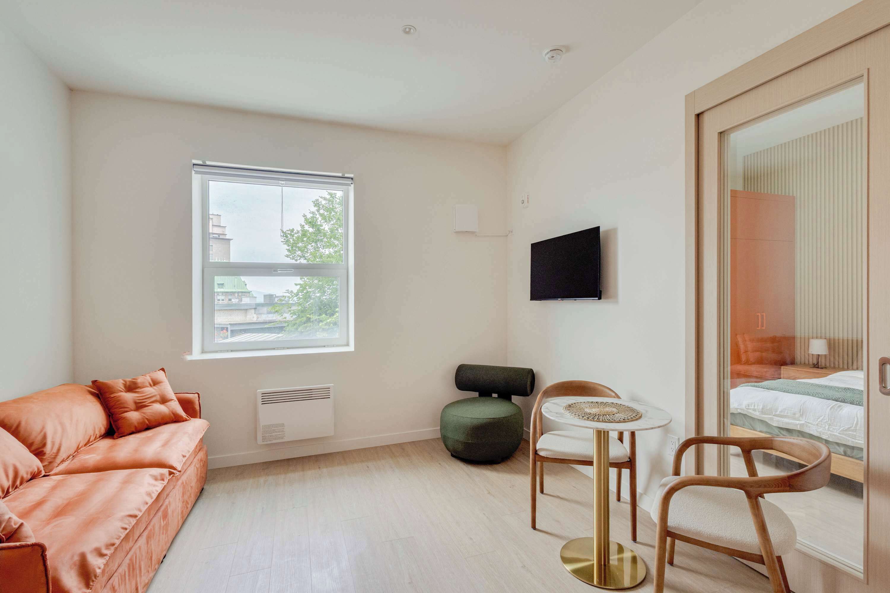 A modern room with a sofa, a small table with chairs, a bed, and a window with blinds.