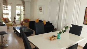 Ein modernes Wohnzimmer mit Essbereich, inklusive weißem Tisch, schwarzen Stühlen und einem Sofa mit bunten Kissen.