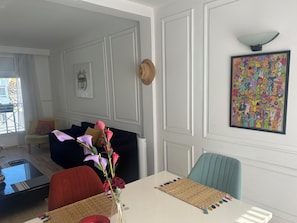 A living room with a sofa, a dining table with a vase of flowers, a painting on the wall, and a window with curtains.