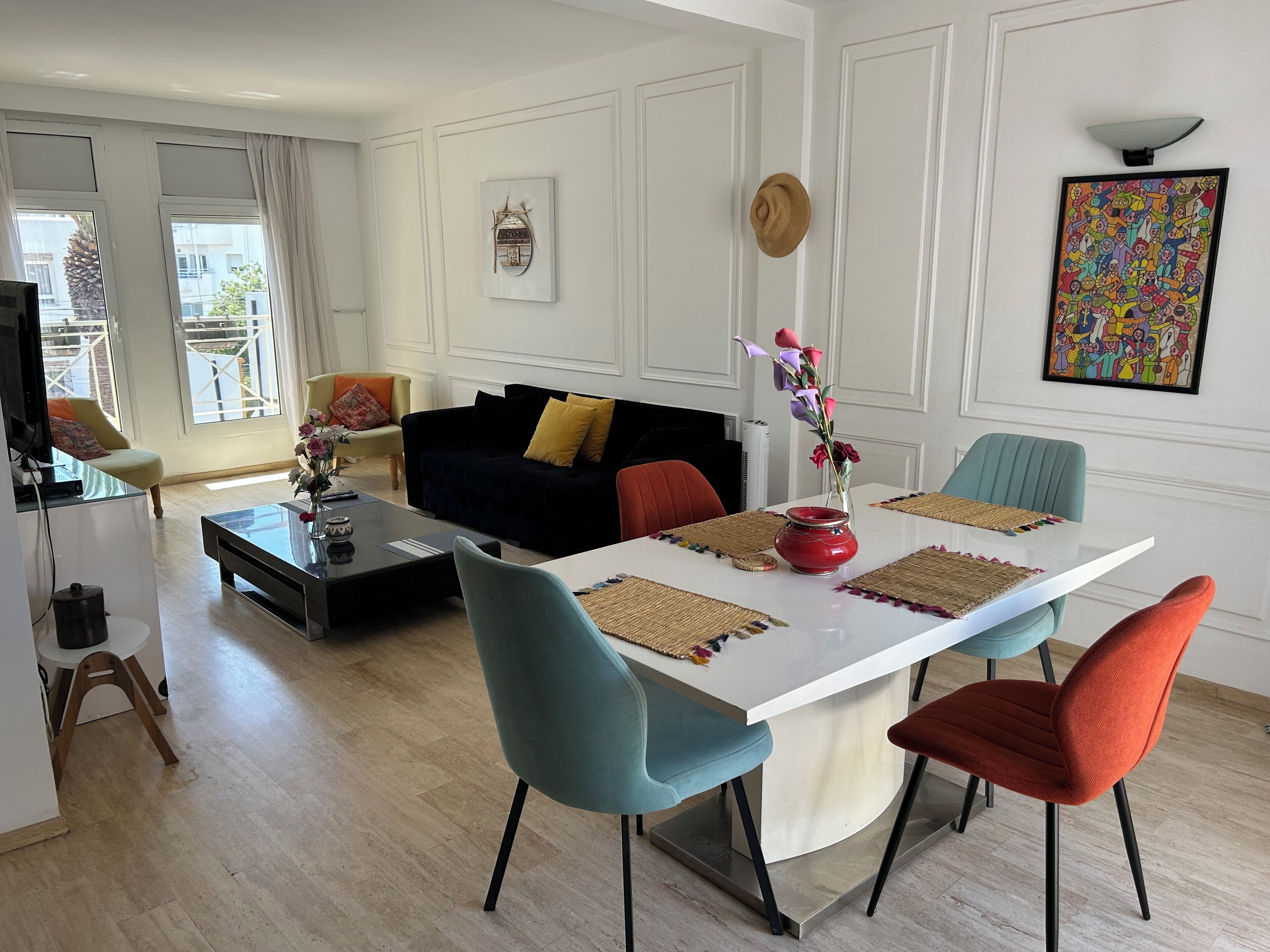 Una sala de estar moderna con una mesa de comedor blanca, sillas coloridas, un sofá negro y un televisor.