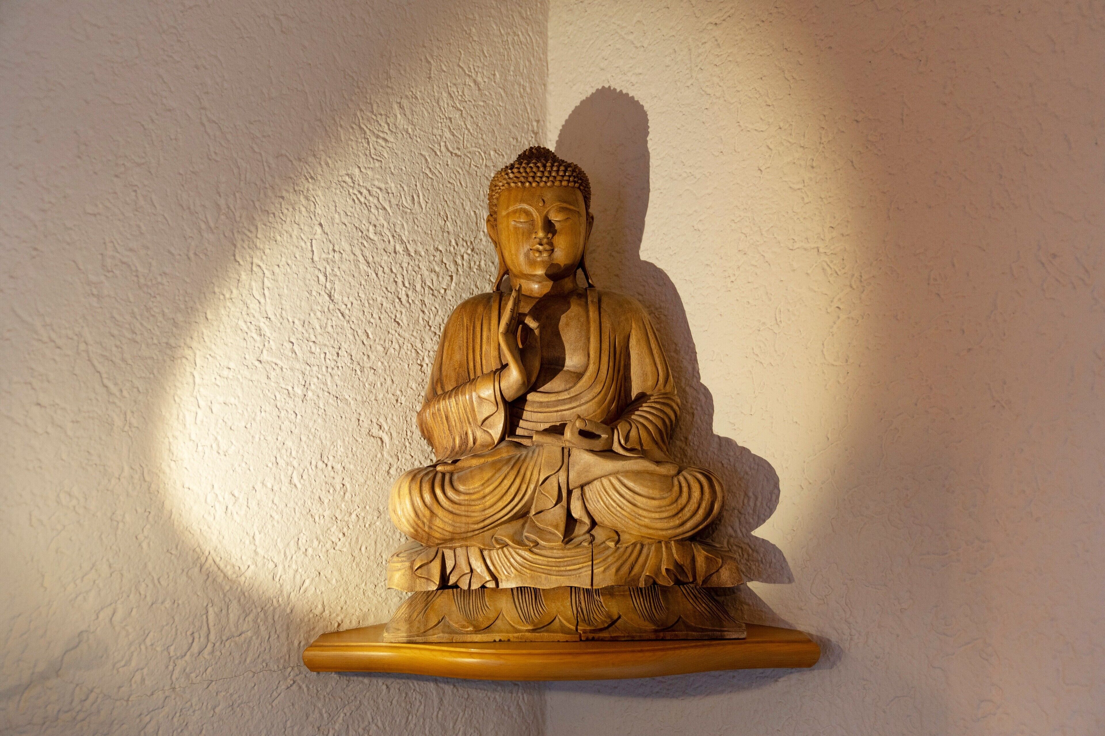 Eine hölzerne Statue einer sitzenden Figur, vermutlich Buddha, auf einem Regal vor einer strukturierten Wand.