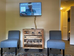 A room with a television mounted on the wall displaying a music album cover, two grey chairs, and a wooden cabinet with metal handles. - Island Street Loft - downtown convenience with proximity to local attractions! (Bryson City)