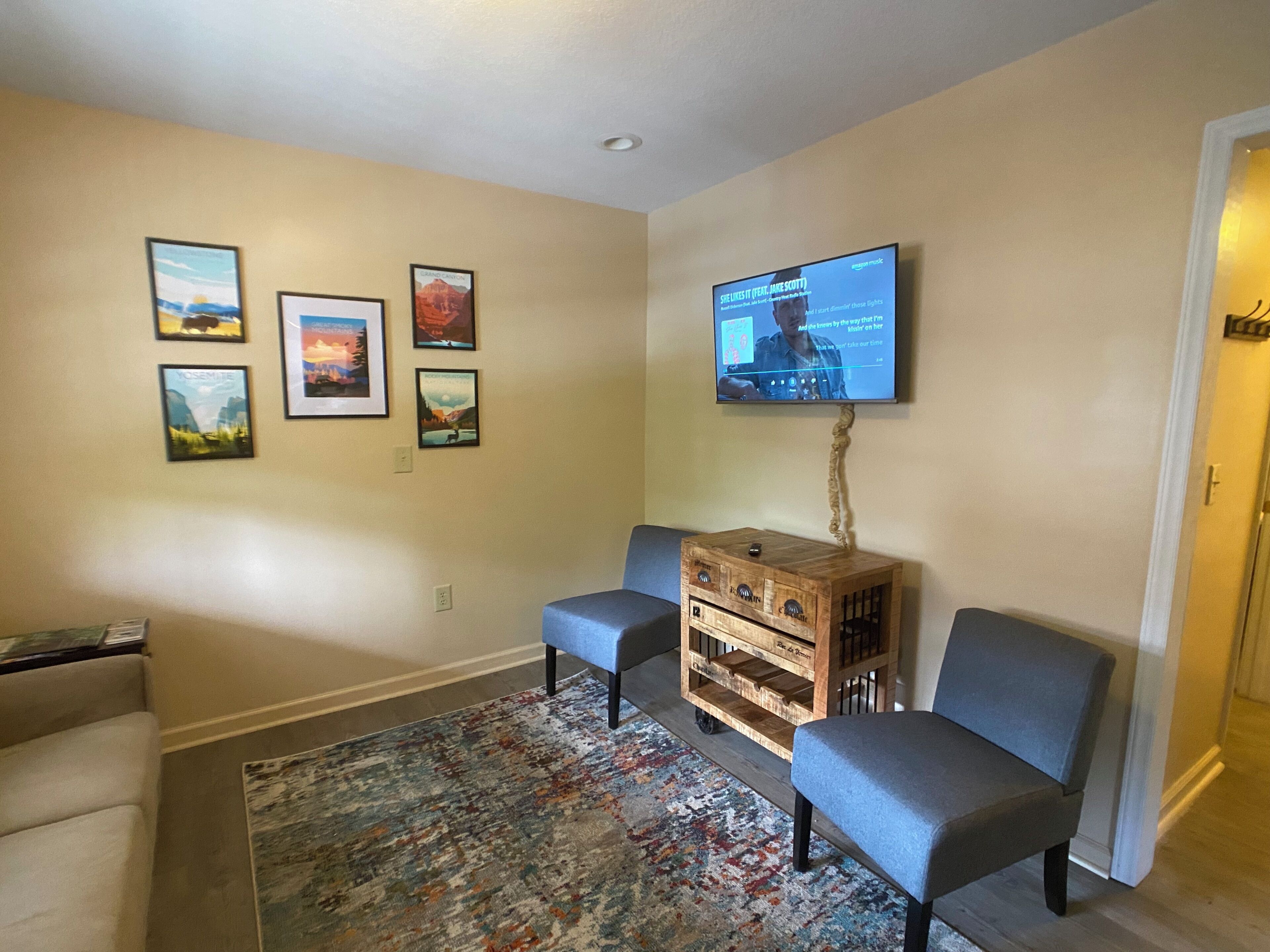 Un salon avec un téléviseur fixé au mur, un tapis à motifs, deux fauteuils et un buffet en bois.