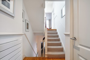 A staircase with a beige carpet and white walls, leading to a room with a window and a door. - Spacious Chelsea Apartment | 3 Bedrooms | Roof Terrace (London)