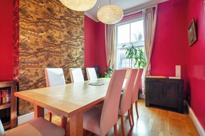 A dining area with a wooden table, white chairs, a stone wall with a mural, a window with curtains, and a plant. - Spacious Chelsea Apartment | 3 Bedrooms | Roof Terrace (London)