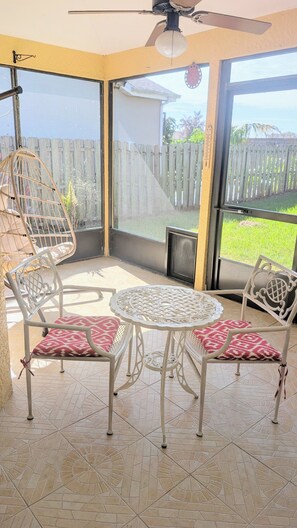 A covered patio area with a table and two chairs, a hanging chair, and a view of a wooden fence and greenery. - Viera Home 3bed 2bath only 5min to Zoo pet-friendly (Melbourne)