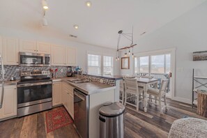 A modern kitchen with stainless steel appliances, a central island, and a dining area with a view. - JL #061 E Z Livin - Hot Tub - Close to beach (Kill Devil Hills)