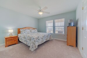 Une chambre à coucher comprenant un lit, une table de chevet, une lampe, un ventilateur de plafond, des stores et une commode.