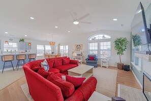 A spacious living area with a red sectional sofa, a dining table with chairs, a kitchen with blue cabinets, and a large window with a view. - JL #313 Sea Bass - Hot Tub - Game Room - Close to Beach (Kill Devil Hills)