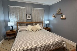 A bedroom with a large bed, two bedside tables with lamps, a headboard, and a wall-mounted shelf with decorative items. - SAGE Boutique Retreat • Sleeps 6 (Burrillville)