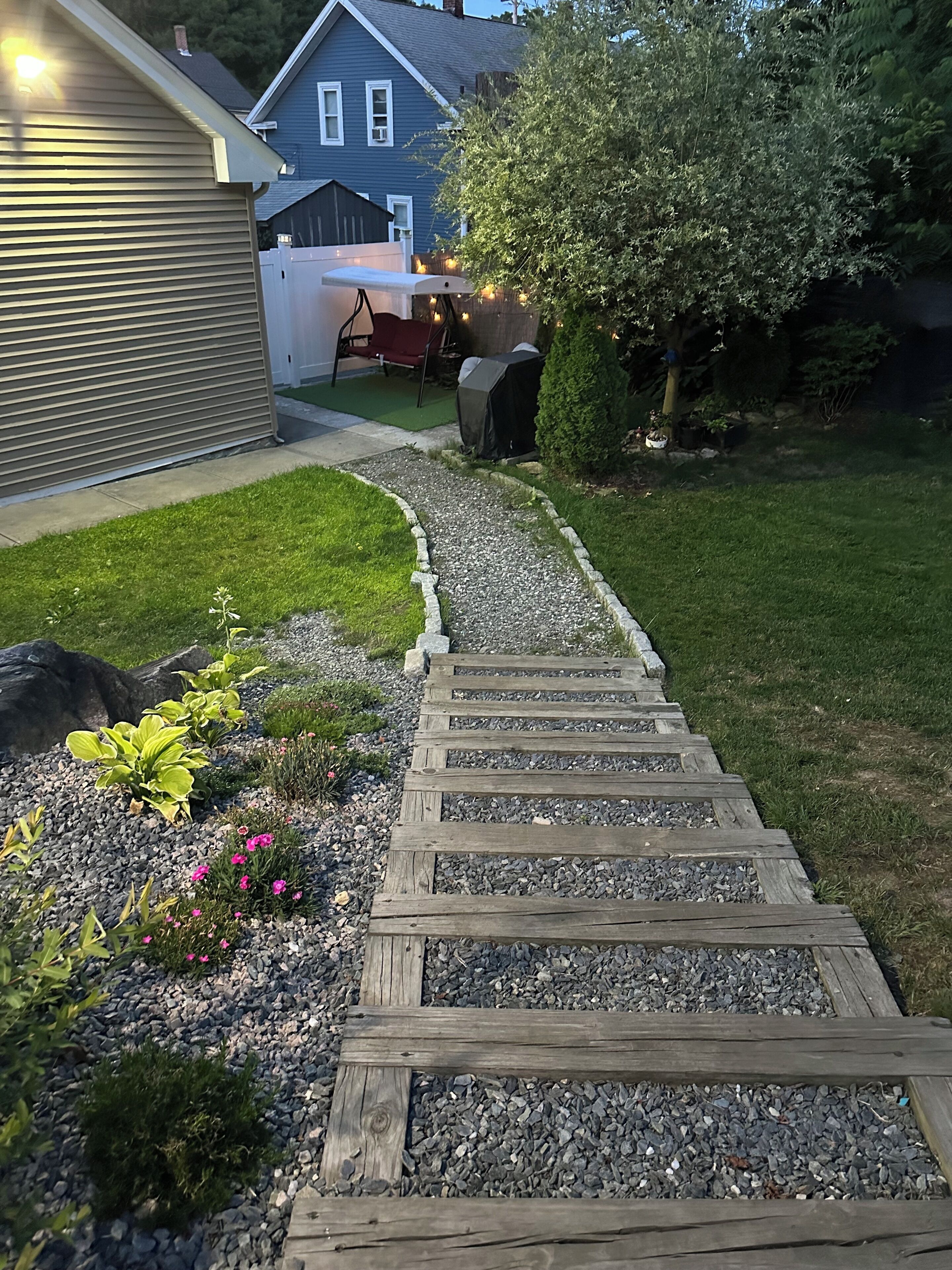 A wooden pathway leading to a house with a garage, surrounded by a well-maintained lawn and garden.