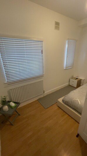 A bedroom with a bed, a bedside table, a radiator, and two windows with blinds. - Luxury 2-Bed London Flat | Free Parking, PS5 & Sleeps 6 (London)