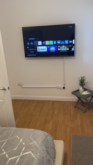 Ein an der Wand befestigter Flachbildfernseher mit Kabelmanagement, ein Nachttisch mit Pflanze und ein Bett mit gemustertem Überwurf.
