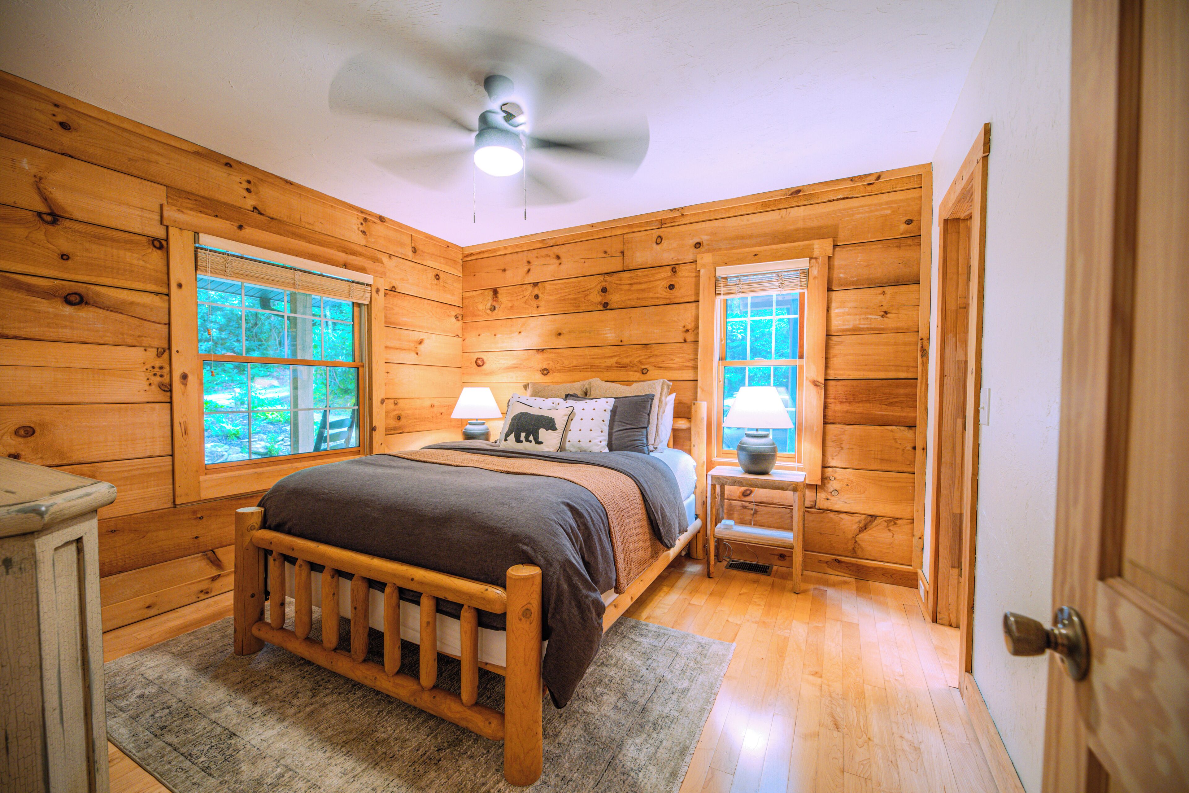 A wooden cabin bedroom with a bed, two bedside tables, a ceiling fan, and two windows.