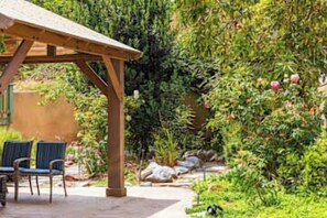 A covered outdoor seating area with chairs and a table, surrounded by lush greenery and flowering plants. - Bay Park Vacation House (San Diego)