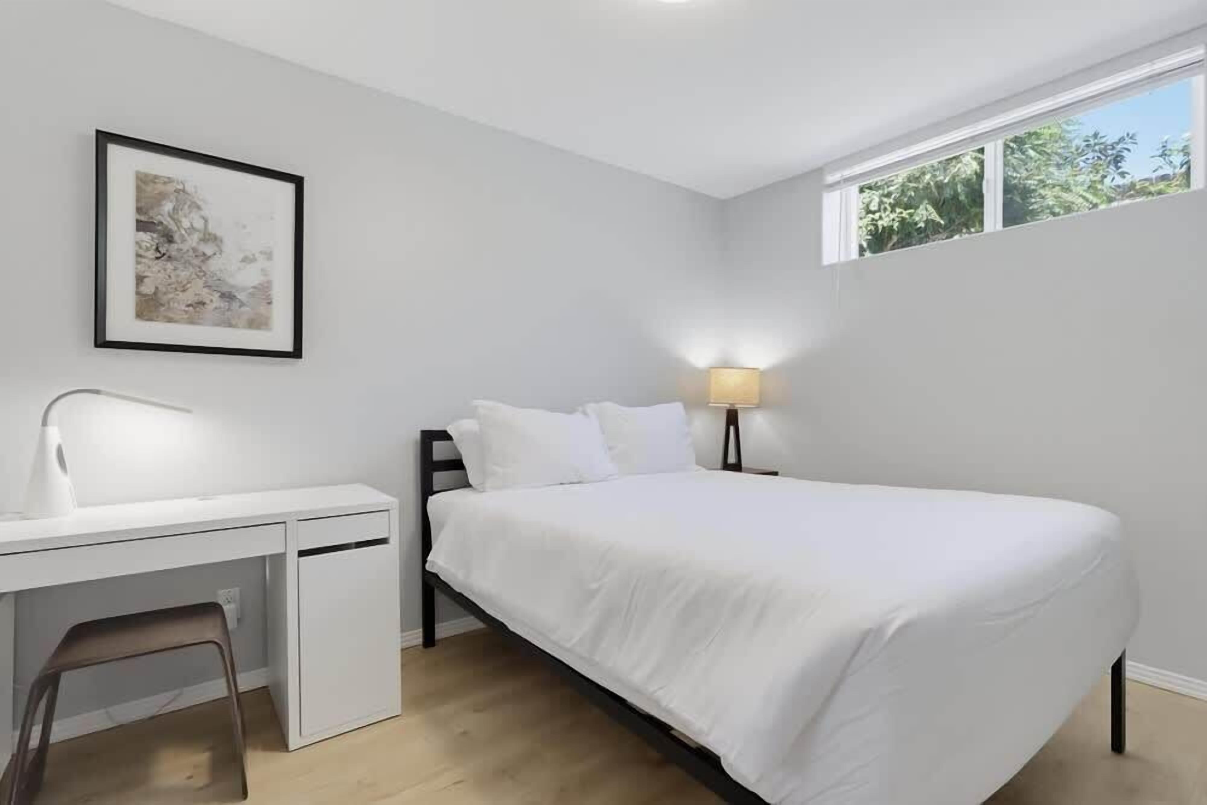 Un dormitorio con cama, escritorio, silla, mesita de noche con lámpara y ventana.