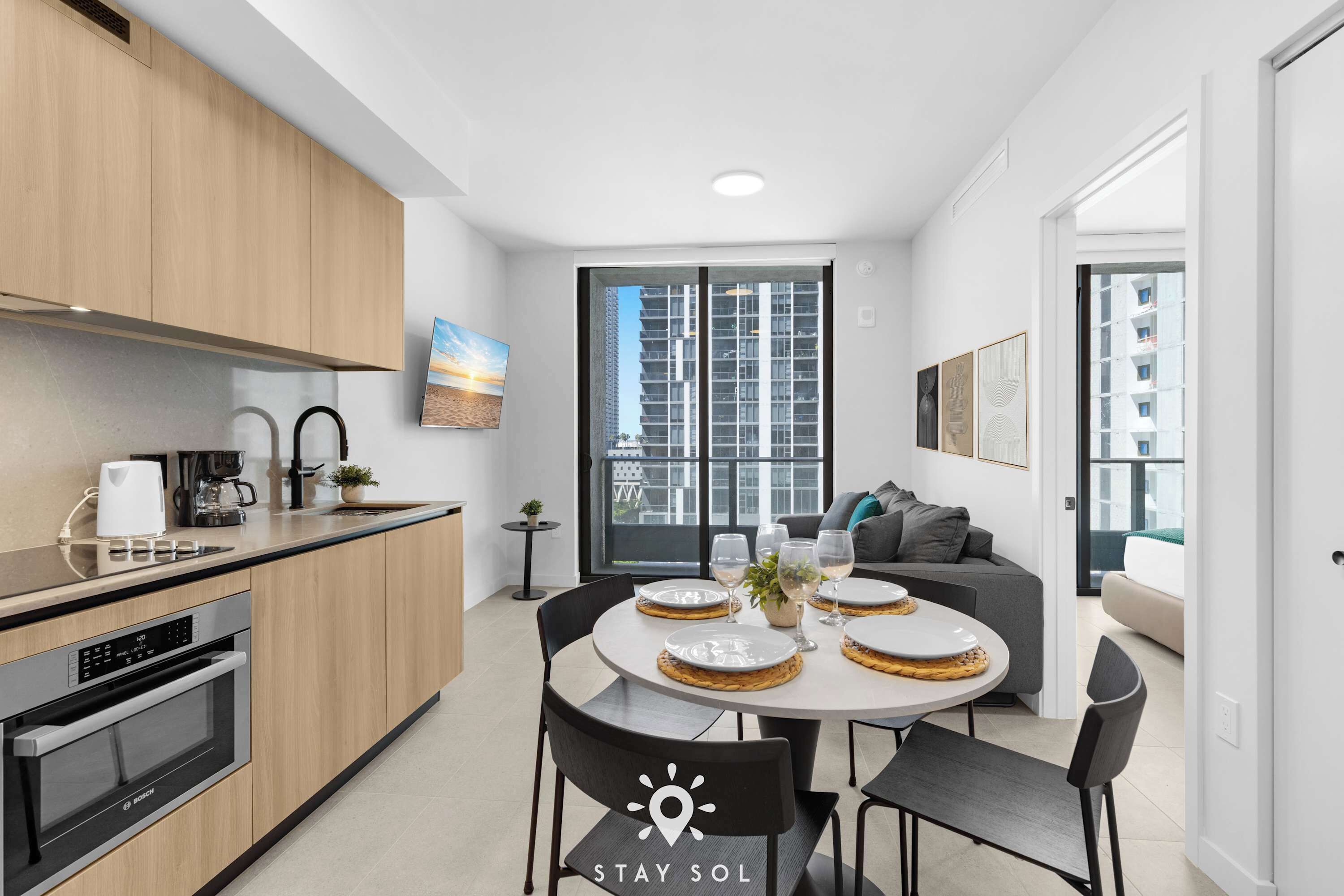 A modern kitchen with built-in appliances, a dining area with a round table and chairs, and a large window offering a view of high-rise buildings.