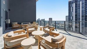 Een dakterras met ronde tafels en houten stoelen, uitzicht over de stad, en een modern gebouw op de achtergrond.