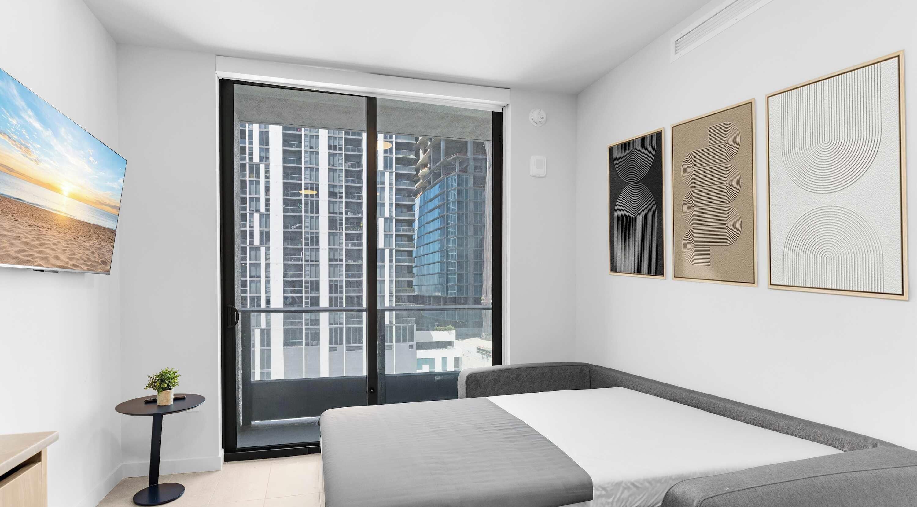 A modern bedroom with a bed, a small table with a plant, a large window with a view of buildings, and three framed artworks on the wall.