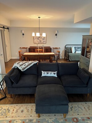 A living room with a dark-colored sofa, a matching ottoman, a dining table with chairs, and a chandelier.