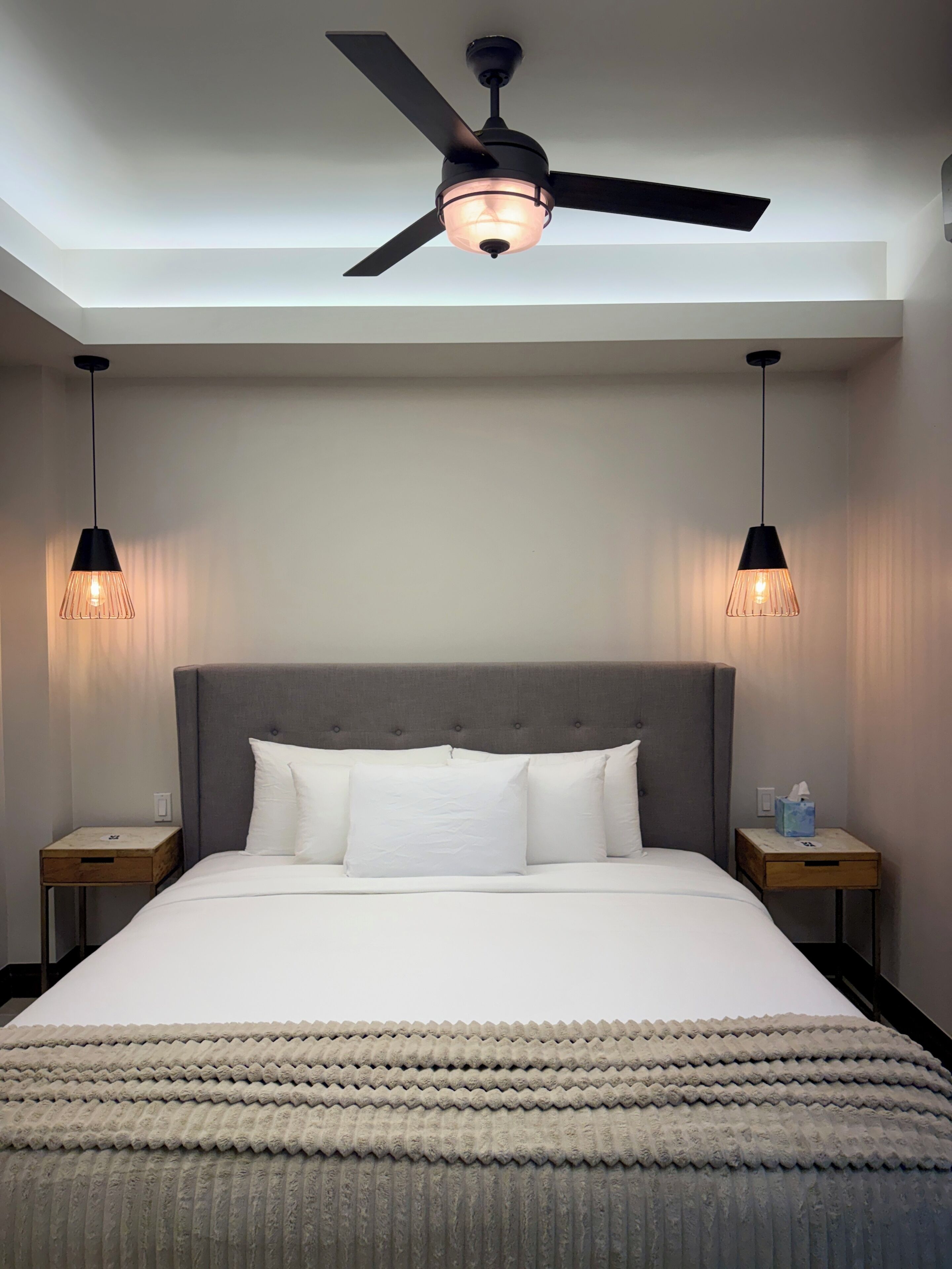 Una habitación de hotel con una cama grande, un ventilador de techo, dos lámparas de noche y una mesita de noche con una caja de pañuelos.