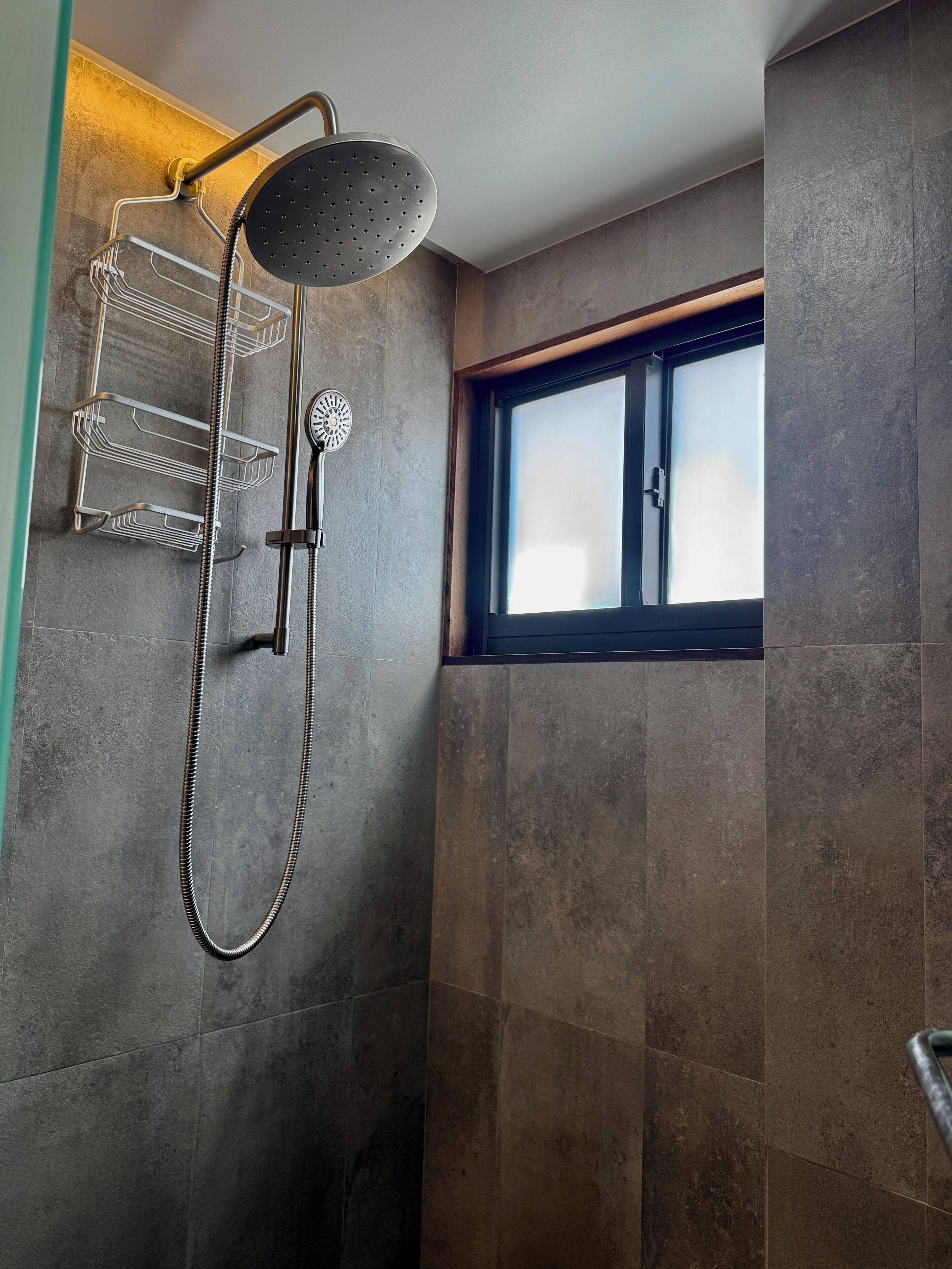 Ducha con cabezal de ducha manual y llave de pared, ventana con persianas y estante metálico con ganchos.