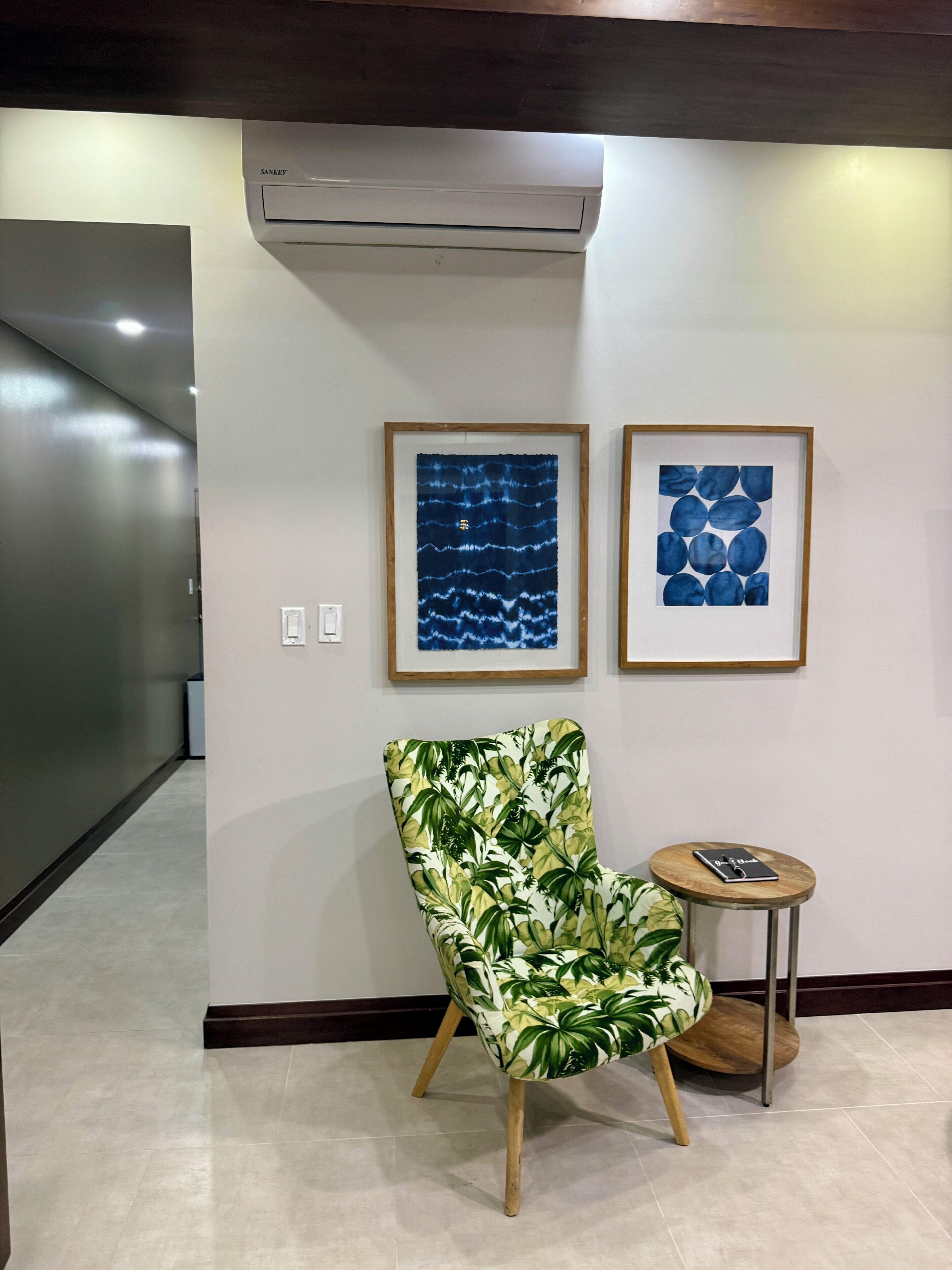 A hallway with an air conditioning unit, a chair with a tropical print, and two framed artworks on the wall.
