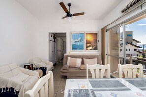Ein gemütliches Wohnzimmer mit einer Couch, Sesseln und einem Esstisch. Es gibt einen Deckenventilator, ein Fenster mit Blick auf Gebäude und ein Bild an der Wand.