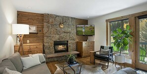 A modern living room with a stone accent wall, a fireplace, a television, a sofa, a coffee table, and a large window with a view. - Remodeled  East Vail Condo w/ Creekside Views, Free Shuttle Access!! (Vail)