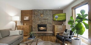 A living room with a stone fireplace, a flat-screen TV, a sofa, a coffee table, and a large plant. - Remodeled  East Vail Condo w/ Creekside Views, Free Shuttle Access!! (Vail)