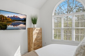 A bedroom with a bed, a wooden dresser, a potted plant, a wall-mounted TV, and a window with blinds.
