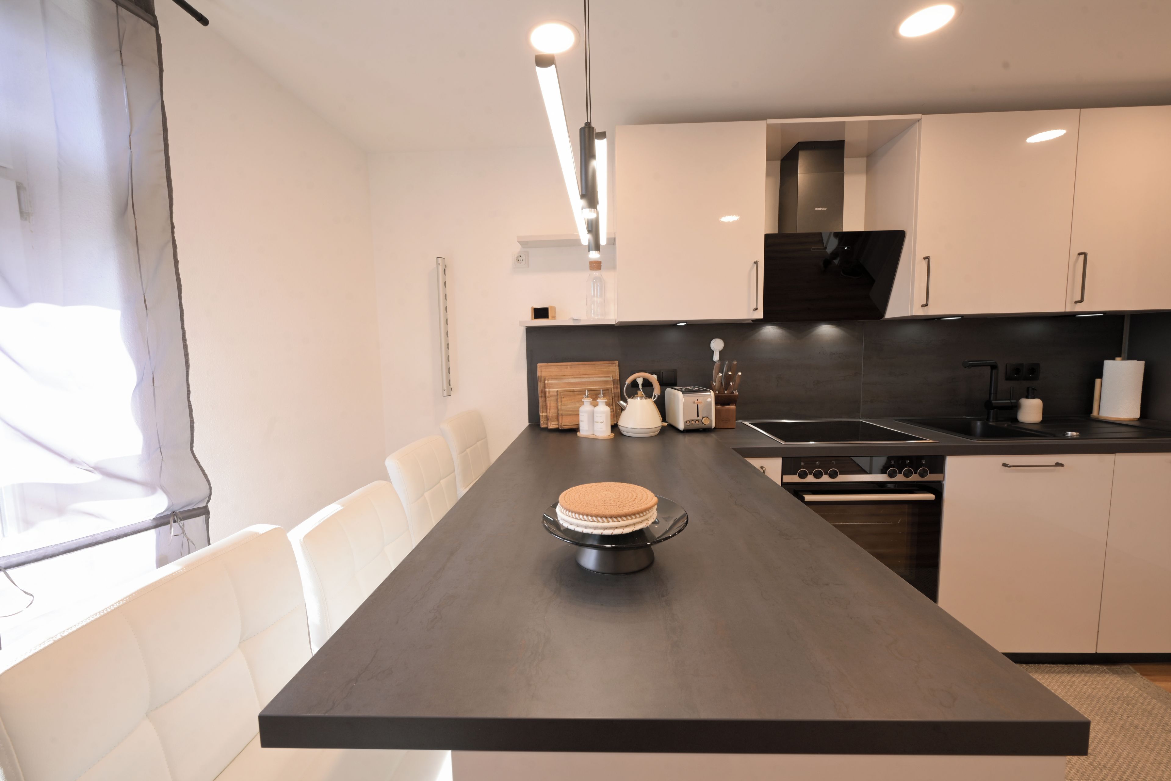A modern kitchen with a dark countertop, white chairs, and a cake on a stand.