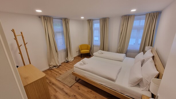 A hotel room with a wooden floor, two beds, a chair, and a coat rack. - LIBORIA Alpine Getaway: Stunning Vacation Rental with a Balcony (Riegsee)