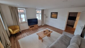 A living room with a sofa, a yellow chair, a TV on a wooden stand, a painting on the wall, and a door leading to a kitchen.