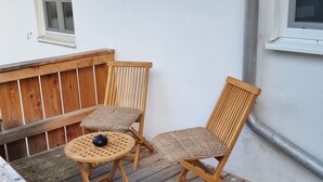 A wooden deck with two chairs and a table, a window, and a white wall with a pipe. - LIBORIA Alpine Getaway: Stunning Vacation Rental with a Balcony (Riegsee)