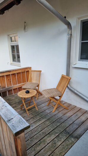 Puisesta laudasta rakennettu terassi, jossa on kaksi tuolia ja pöytä, ikkuna sekä valkoinen seinä, jossa kulkee putki.