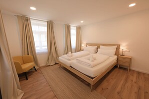 Quarto de hotel com cama de madeira, uma cadeira, mesa de cabeceira e janela com cortinas.