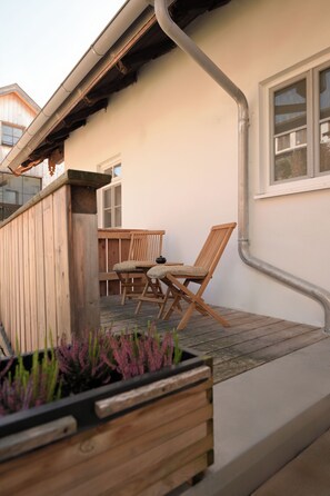A wooden deck with a table and chairs, a planter with plants, and a building with a window and a door.