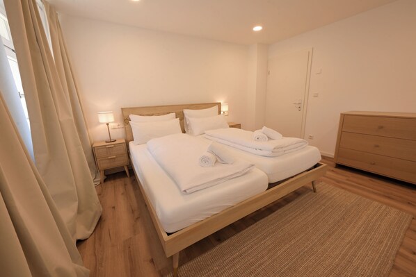 A double bed with white linens, a bedside table with a lamp, a wooden dresser, and a door.