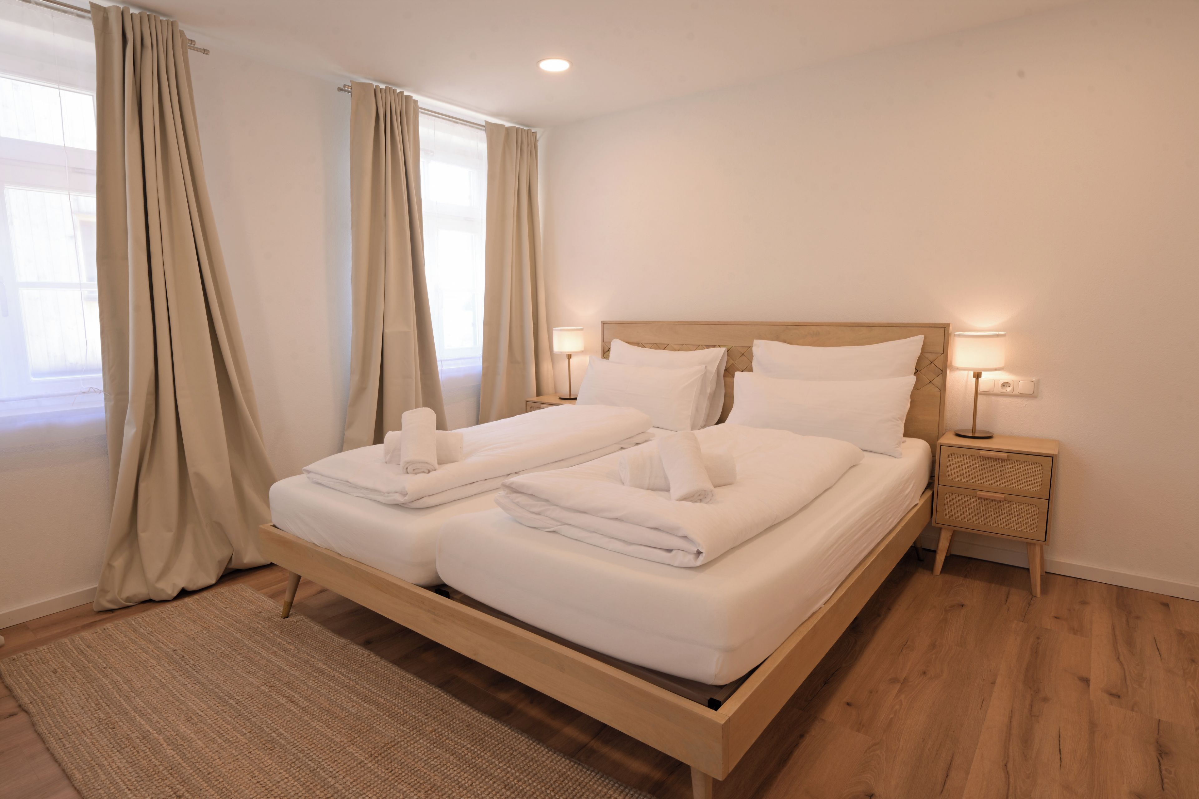 Un lit double avec du linge de lit et des serviettes blanches, une tête de lit en bois, des lampes de chevet et un tapis tissé sur un plancher en bois.