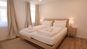 Un lit double avec du linge de lit et des serviettes blanches, une tête de lit en bois, des lampes de chevet et un tapis tissé sur un plancher en bois.